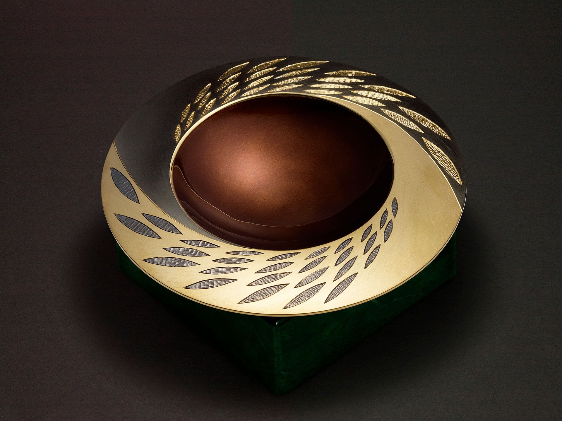 Angle view: ELEMENTS SPINNING BOWL - EARTH 2007 reworked 2009. 150mm diameter. Silver, hand engraved / carved, black and yellow gilding, earth/bronze/brown enamel. Base in green verdite. Photo : Simon B Armitt.