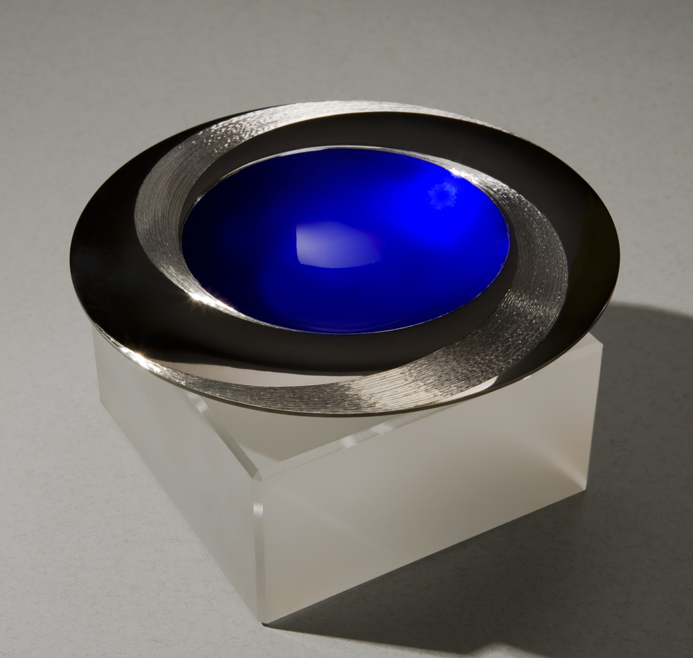 Angle view: BLUE INFINITY BOWL 150 mm diameter. Britannia Silver. Blue Enamel, Black Gilding, Hand Engraved. Base frosted and polished Optical Quartz. Bowl is designed to spin gently on its stone base. Photo : Simon Armitt